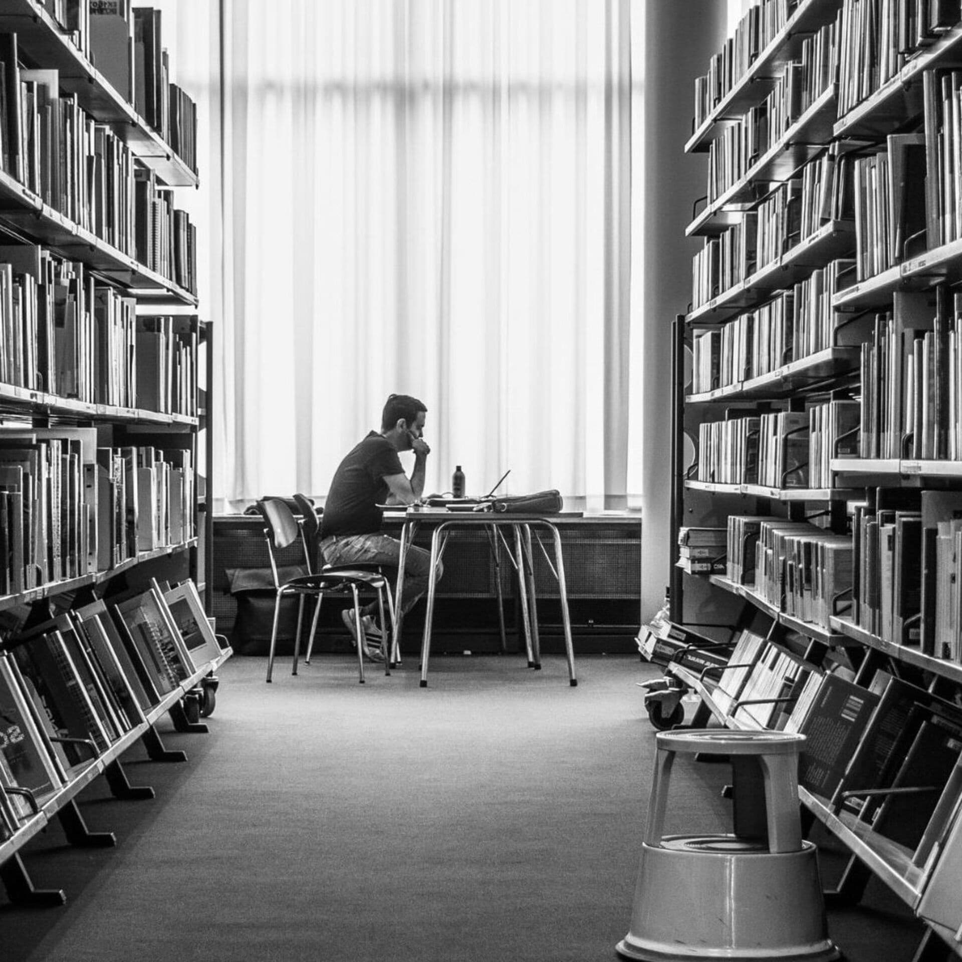 En person sitter vid ett bord med en laptop mellan två bokhyllor i ett bibliotek.