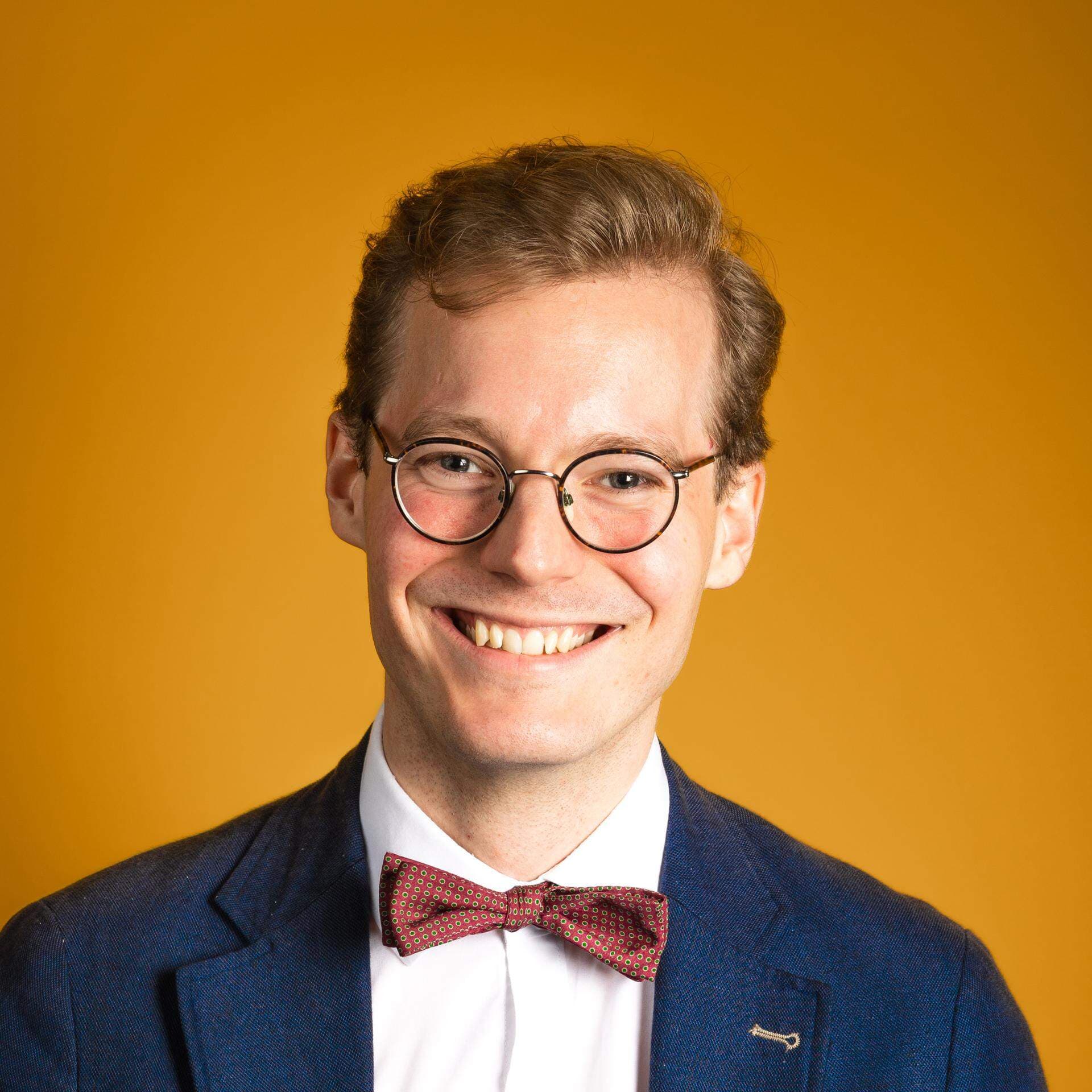 Caspian Rehbinder iklädd vit skjorta, mörkblå kavaj och röd fluga. Bakgrunden är orange