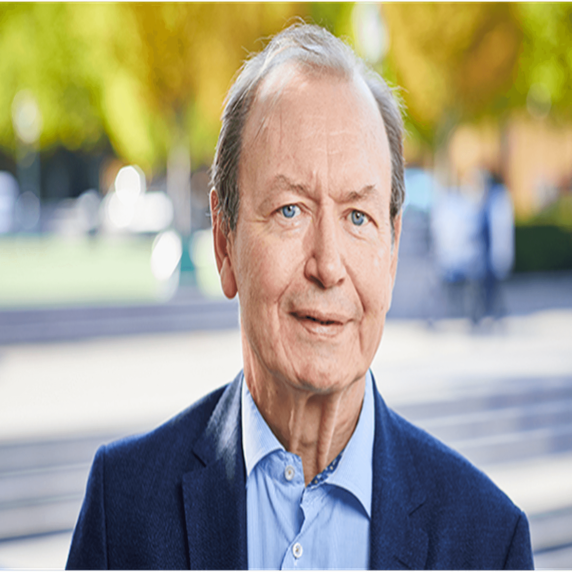 Gert Nilsson iklädd blå skjorta och blå kavaj, fotograferad utomhus i stadsmiljö med gröna träd i bakgrunden