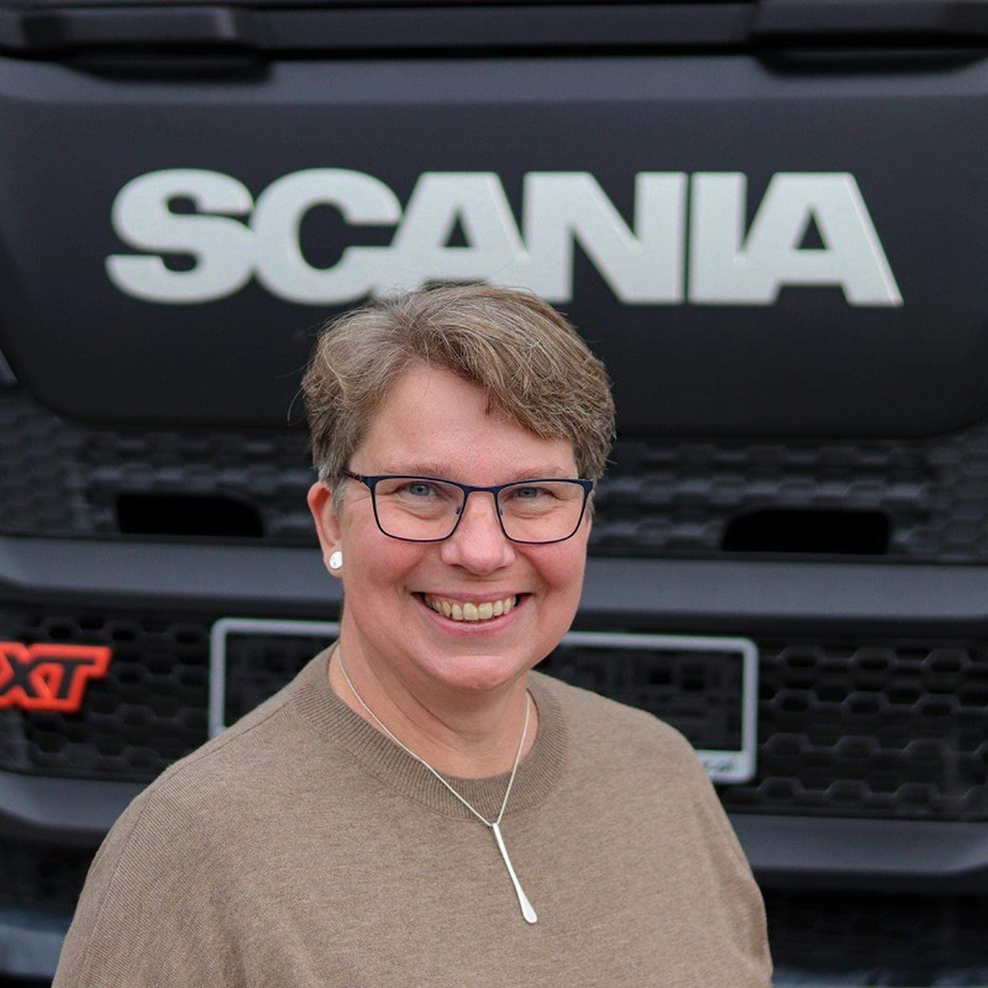 Anna Olshäll iklädd brun stickad tröja med en lastbilsfront i bakgrunden