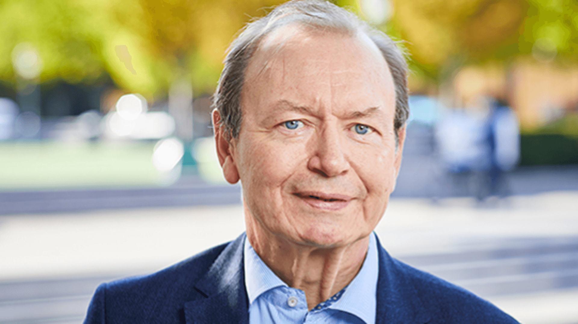 Gert Nilsson iklädd blå skjorta och blå kavaj, fotograferad utomhus i stadsmiljö med gröna träd i bakgrunden