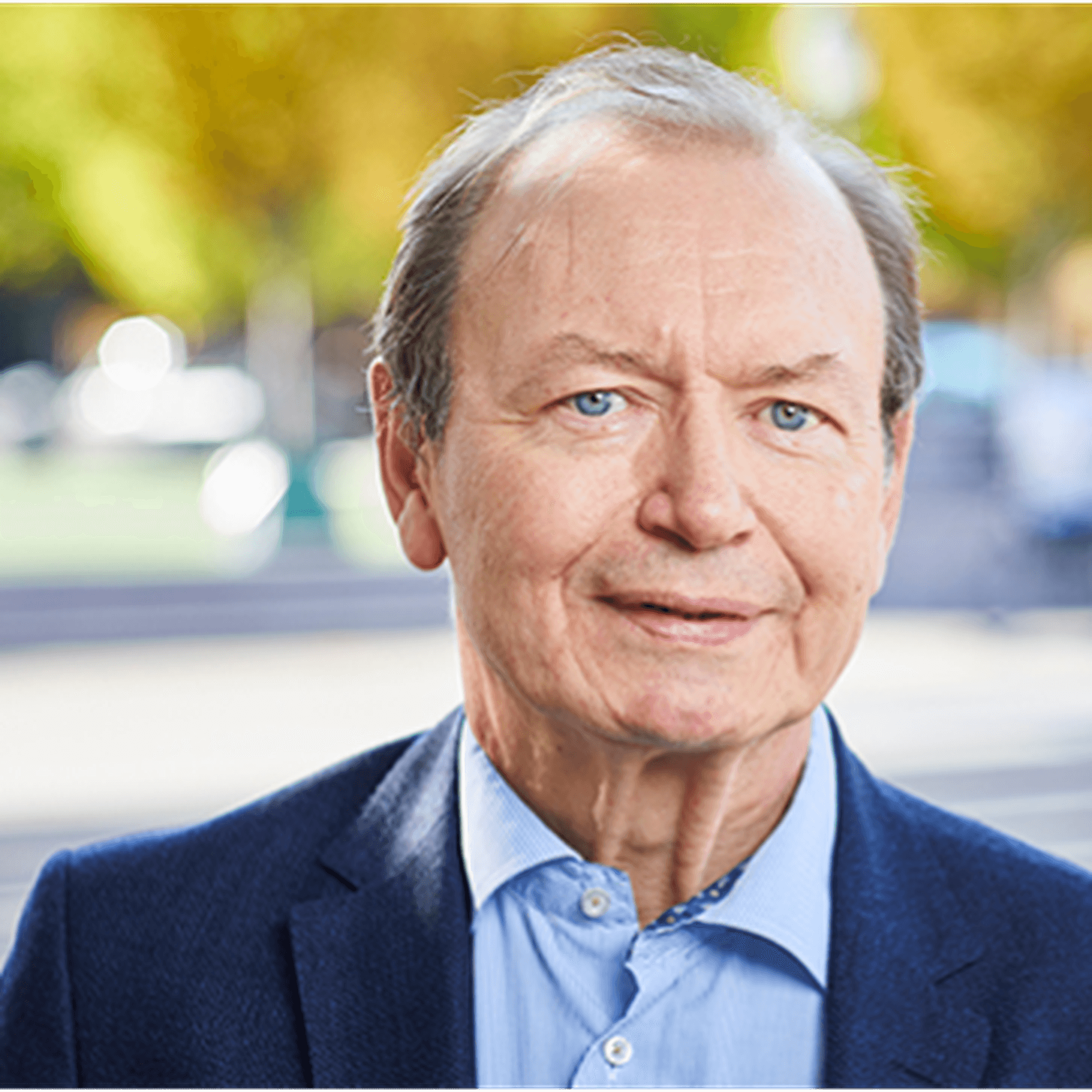 Gert Nilsson iklädd blå skjorta och blå kavaj, fotograferad utomhus i stadsmiljö med gröna träd i bakgrunden