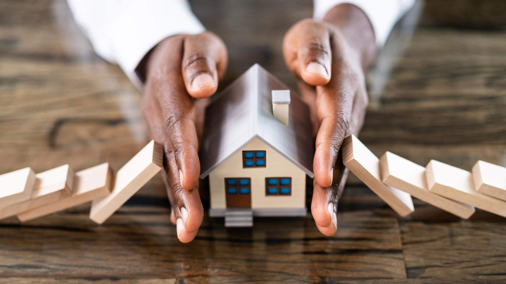 Händer som skyddar ett leksakshus från brickor som lutar emot.