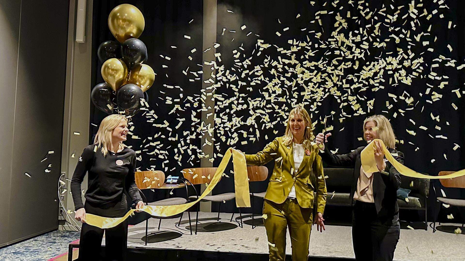 Tre personer håller i ett guldband och det är ballonger och confettiregn